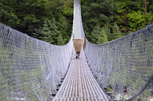 A person carrying a woven basket across a suspension bridge surrounded by green forest